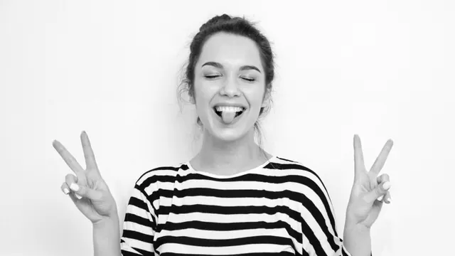 Photo noir et blanc d'une femme souriante, langue tirée, yeux fermés, signe peace des deux mains, pull rayé